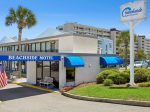 Beachside Motel exterior with blue awnings, a white facade, palm trees, and a tall Beachside Motel sign in a sunny coastal setting.